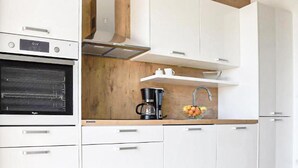 Une cuisine moderne avec des meubles blancs, un four encastré, une machine à café et un panier à fruits posé sur le plan de travail.