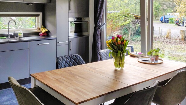 Une cuisine moderne avec des meubles gris, une table à manger en bois et une vue sur le jardin par la fenêtre.