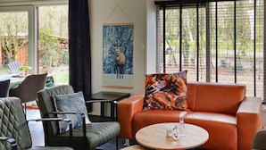 Un salon moderne avec un canapé en cuir, des fauteuils et une table basse. Un tableau représentant un cerf est accroché au mur, et la pièce est dotée de grandes fenêtres équipées de stores.