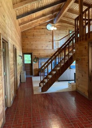 A wooden interior with a staircase leading to a second floor, a ceiling fan, and a brick floor.