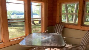 A wooden cabin interior with a glass-top table and striped chairs, large windows offering a view of a lake and trees. - Goose Lake Cabin - two lakes to enjoy! (Spooner)