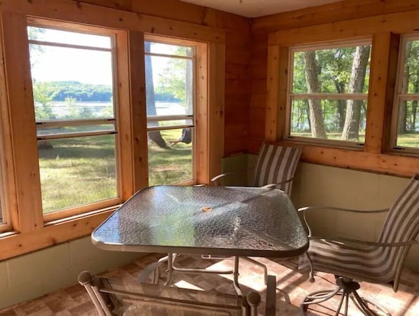 A wooden cabin interior with a glass-top table and striped chairs, large windows offering a view of a lake and trees.