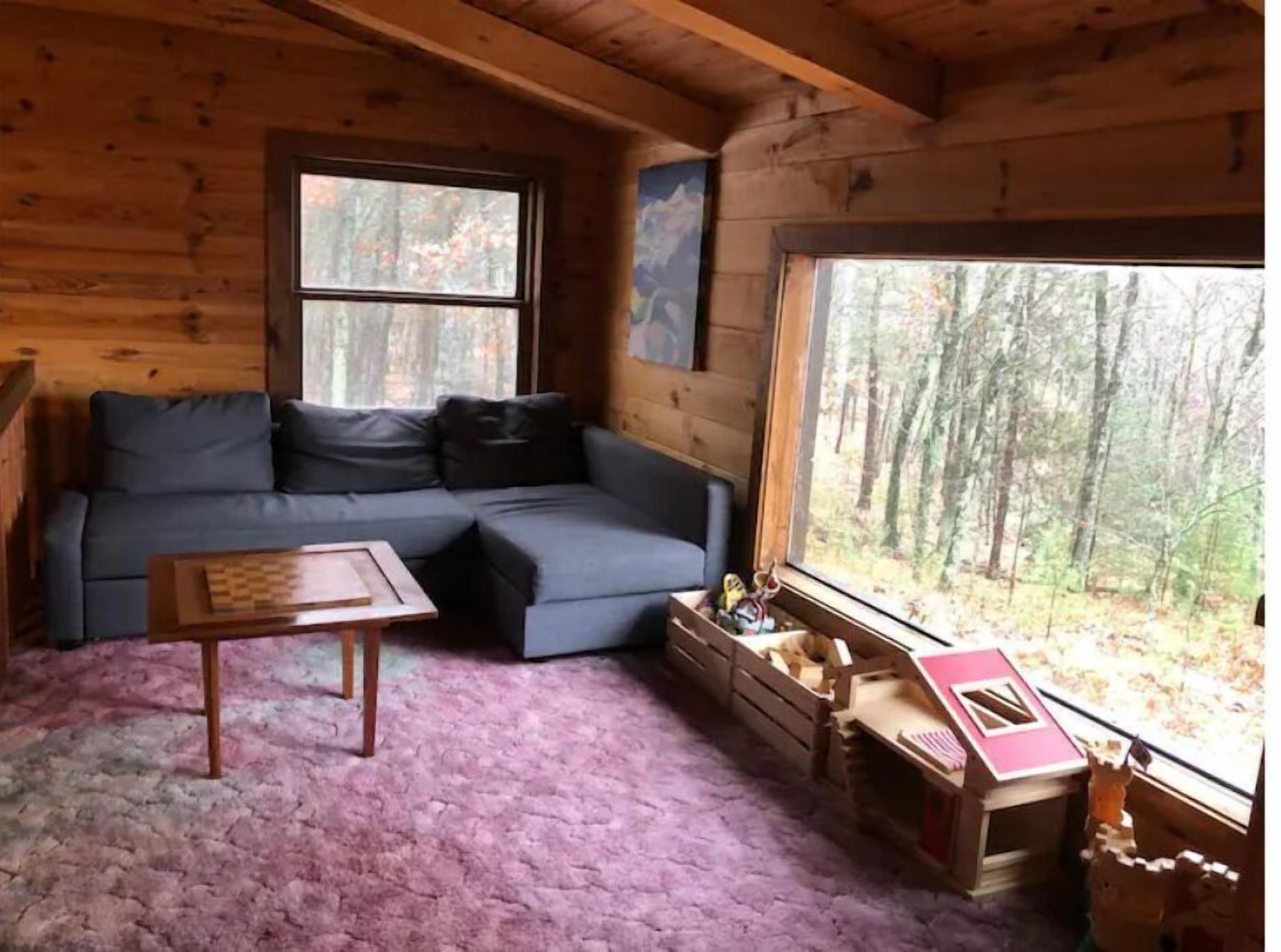 Interior acogedor de una cabaña con un sofá seccional, una pequeña mesa de madera y un ventanal con vista al bosque.