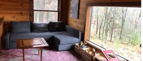 Interior acogedor de una cabaña con un sofá seccional, una pequeña mesa de madera y un ventanal con vista al bosque.
