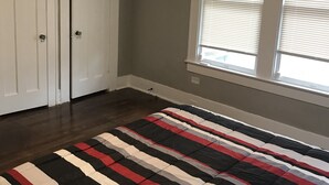 Une chambre à coucher avec une couvre-lit à rayures, une grande fenêtre avec des stores et une porte dotée d’une poignée.