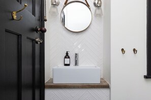 A bathroom with a white sink, a round mirror, and a wooden shelf. - Bristol Gardens - Luxury Home, 2 min from the Beach (Brighton)