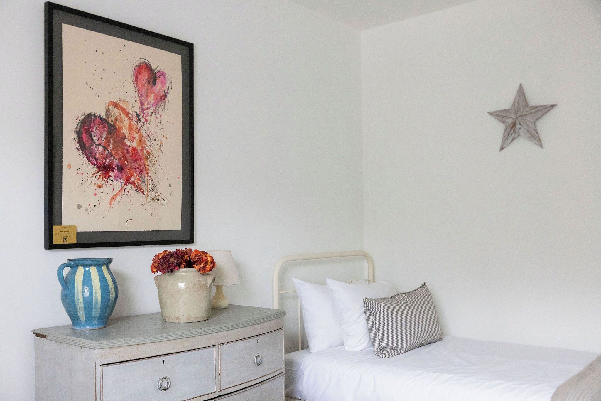 A bedroom with a bed, a dresser, a framed artwork, a vase with flowers, and a star decoration on the wall.