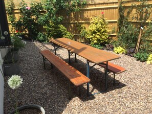 A wooden picnic table with benches in a garden area with gravel ground, surrounded by various plants and a wooden fence. - Bristol Gardens - Luxury Home, 2 min from the Beach (Brighton)