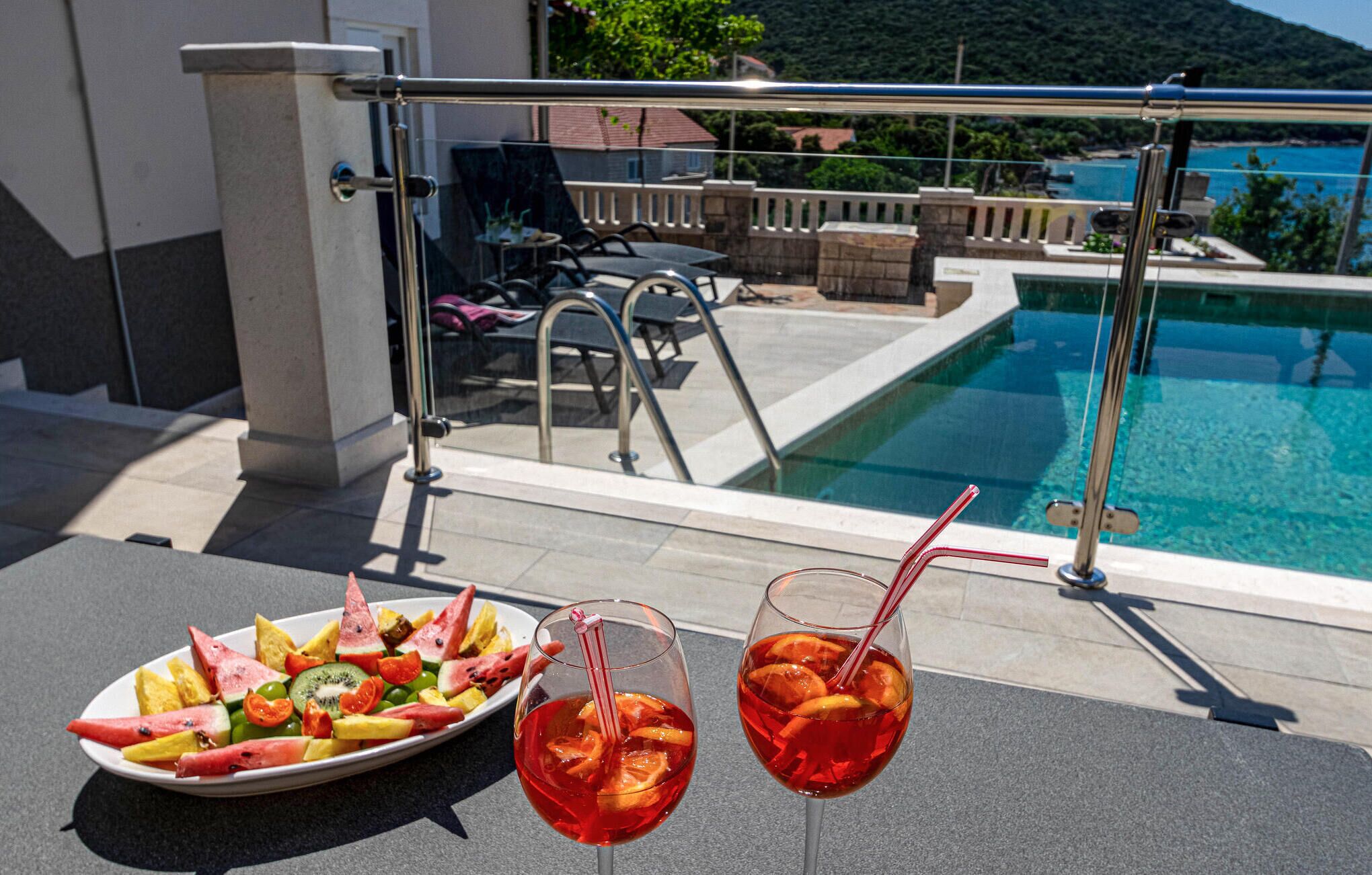 A poolside table with a fruit platter and two drinks, overlooking a pool and a mountainous landscape.