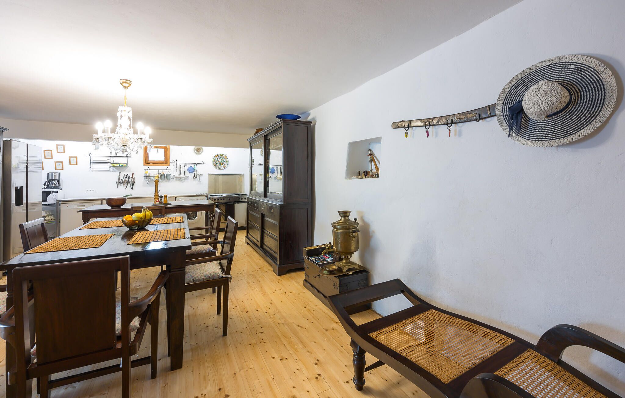 A dining area with a wooden table and chairs, a chandelier, and a kitchen in the background.
