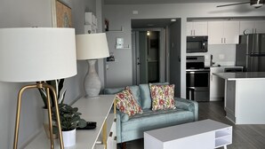 Ett modernt vardagsrum med en soffa, ett soffbord och en lampa. Det finns också en köksdel med vitvaror och skåp.