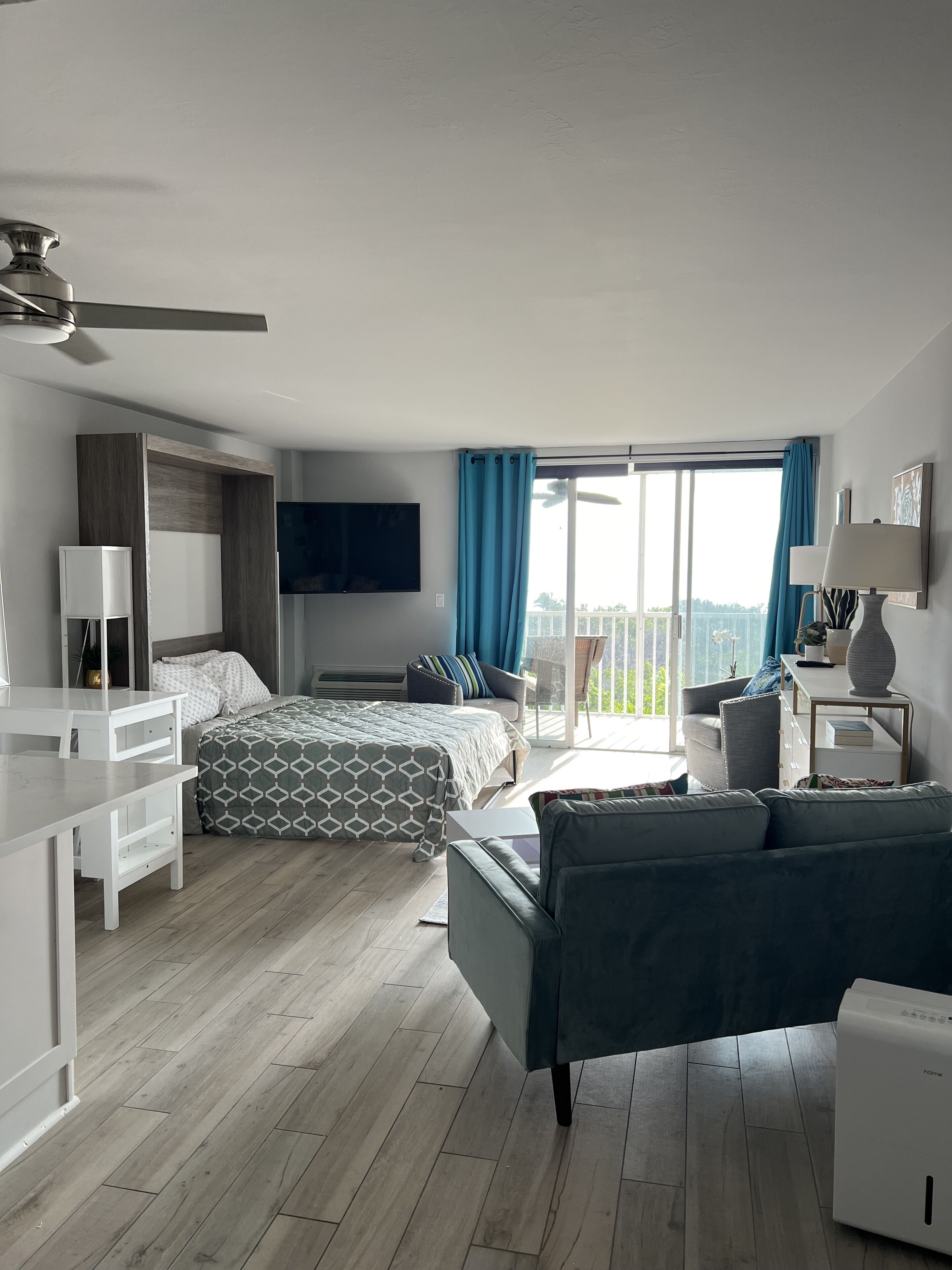A modern hotel room with a bed, a desk, a sofa, a TV, and a balcony with a view.