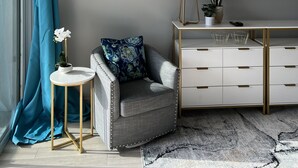 Ett modernt vardagsrum med en grå soffa, ett litet runt bord med en vit blomma, en blå gardin, ett vitt skåp med guldfärgade handtag och en inramad bild av palmer.