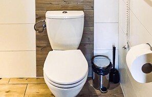 A toilet with a tank, a bidet, and a toilet paper holder in a bathroom with wooden flooring and white walls.