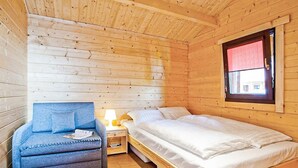 Puinen mökkimäinen huone, jossa on sänky, sininen tuoli, yöpöytä ja ikkuna, jossa on punainen verho.