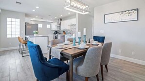 A modern dining area with a wooden table set for four, blue and grey chairs, and a hanging light fixture. - 5BR/3BA NEW Game Room, Pool + Arcade + Fire Pit! (Henderson)
