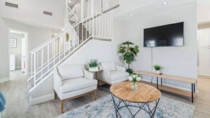 A modern living room with a staircase, a flat-screen TV, a round wooden coffee table, and a white sofa. - 5BR/3BA NEW Game Room, Pool + Arcade + Fire Pit! (Henderson)