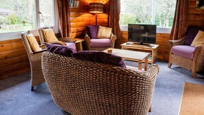 Een gezellig houten blokhut interieur met rieten meubels, een televisie en grote ramen met uitzicht op de omgeving.