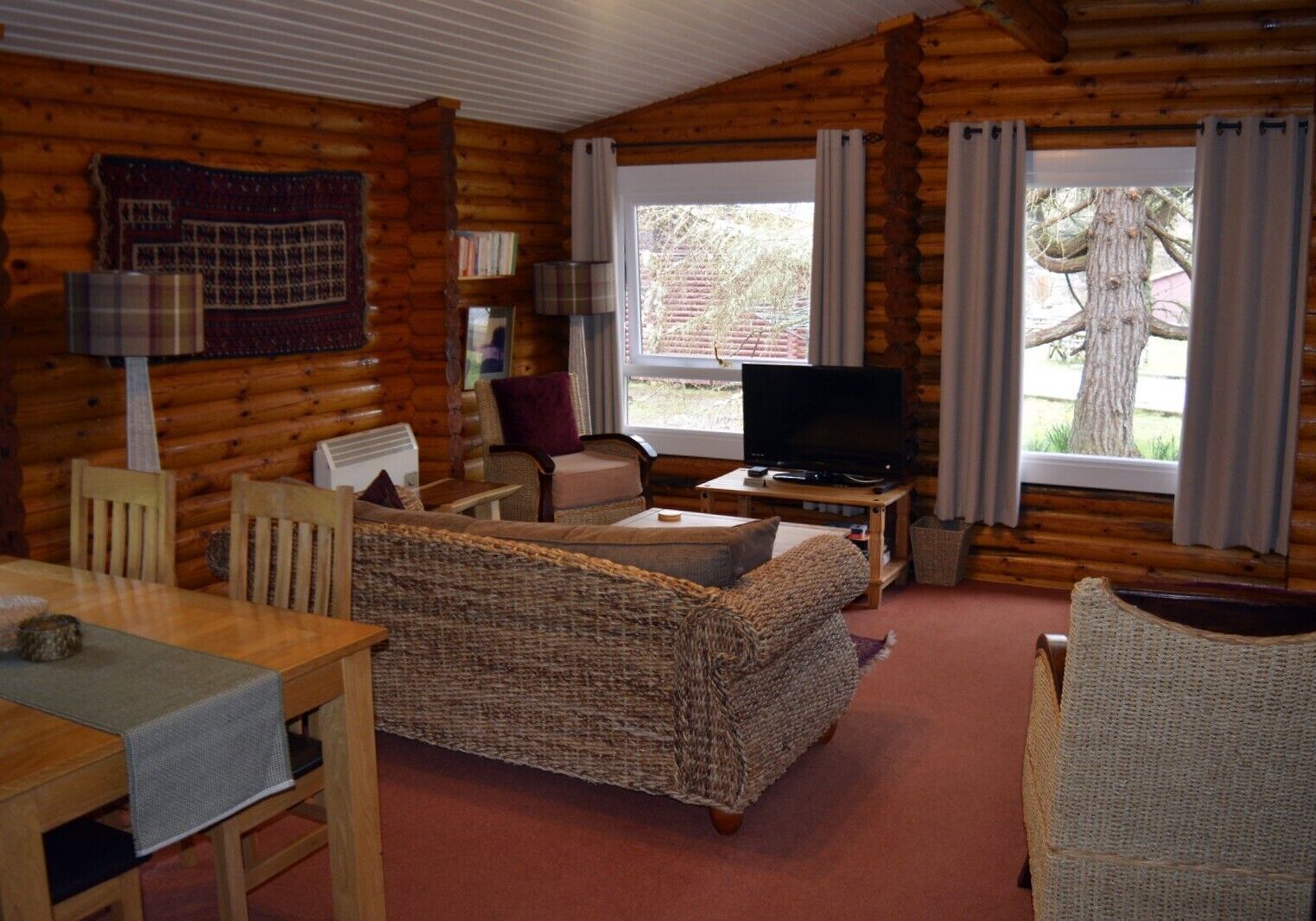 Une pièce confortable, aménagée comme un chalet, avec un intérieur en bois, un téléviseur posé sur un meuble en bois, un canapé en osier et une table à manger avec des chaises.