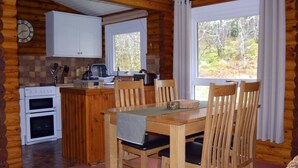 Ein Interieur einer Holzhütte mit einem Holzespeise Tisch, Stühlen, einer Küche mit zwei Öfen, einer Uhr und einem Fenster mit Vorhängen.