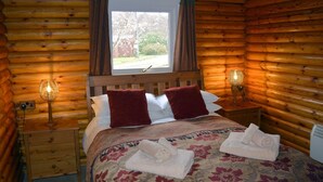 Ein Schlafzimmer in einer Blockhütte mit einem hölzernen Bett, zwei Kissen, einem Fenster und zwei Nachttischlampen.