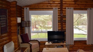 Ein Blockhaushaus-Innenraum mit Fernseher, einem Korbstuhl, einem kleinen Tisch und einem Fenster mit Vorhängen.