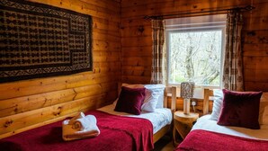 A cozy cabin room with two beds, each with a red blanket and a rolled towel. There's a wooden headboard, a window with curtains, and a wall-mounted lamp.