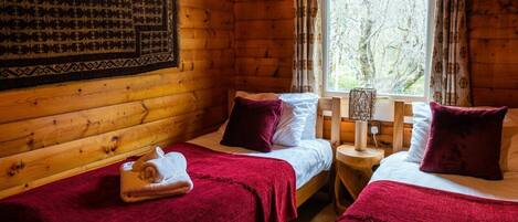 A cozy cabin room with two beds, each with a red blanket and a rolled towel. There's a wooden headboard, a window with curtains, and a wall-mounted lamp.