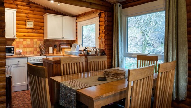 A cozy cabin-style kitchen with wooden walls, a dining table with chairs, a stove, and a window with curtains. - Self-Catering log cabin near Plockton and the Isle of Skye secluded, well equipped and dog friendly. (Kyle)