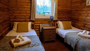 A log cabin room with two beds, a small table, and a window with curtains. - Self-Catering log cabin near Plockton and the Isle of Skye secluded, well equipped and dog friendly. (Kyle)