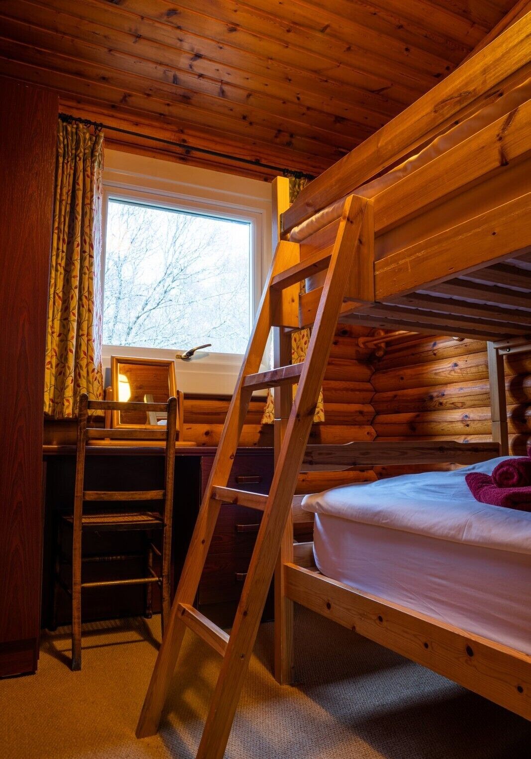 A bunk bed room with a wooden ladder, a desk with a chair, and a window with curtains.