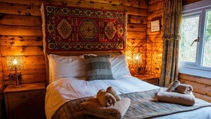 A cozy wooden cabin bedroom with a large bed, two bedside lamps, a patterned headboard, and a window with curtains.