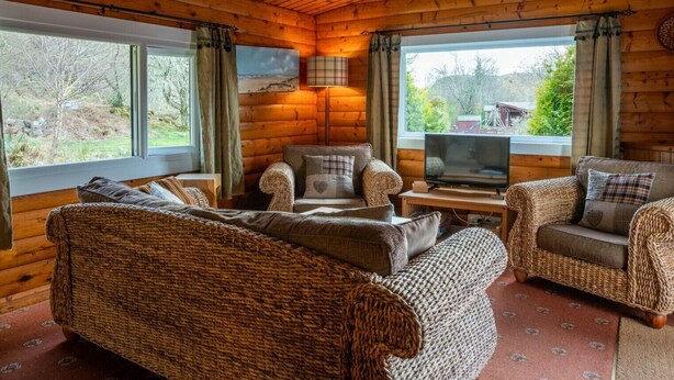 A cozy log cabin interior with a large woven sofa, a smaller sofa, and a television on a wooden table.