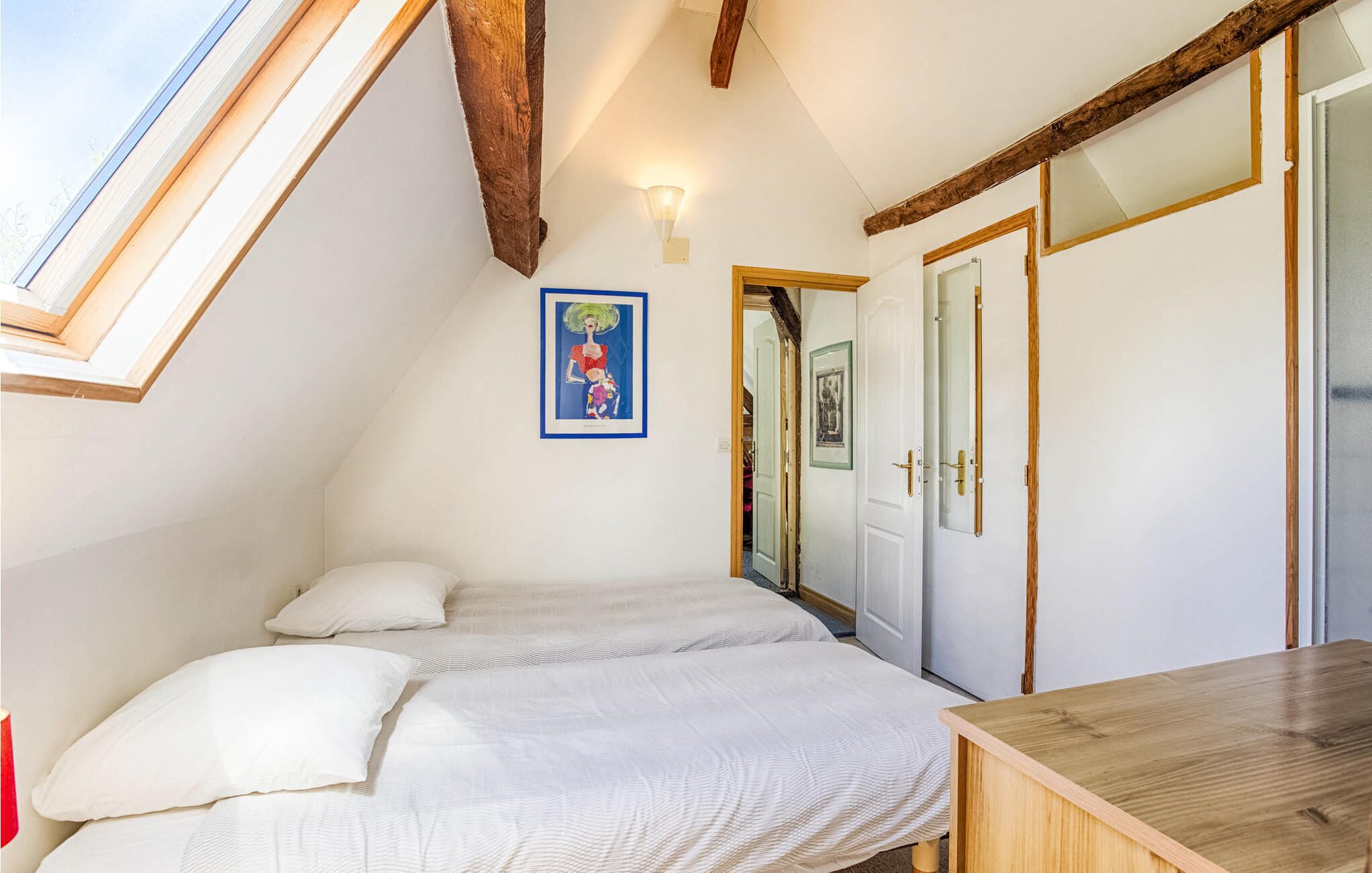 Une chambre avec une lucarne, un lit recouvert de linge de maison blanc, un bureau en bois et un tableau encadré au mur.