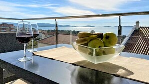 Un balcone con vista libera su un panorama urbano e sull'acqua, un bicchiere di vino rosso e una ciotola di frutta fresca.