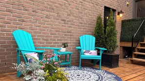 Una terraza de madera con dos sillas Adirondack turquesa, un tapete redondo con estampado y una maceta con flores blanco y rosa.