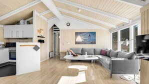 A modern living room with a grey sofa, a dining table with a bowl of fruit, and a kitchen area with white cabinets and a stove. - 4 bedroom beautiful home in Jerup (Jerup)