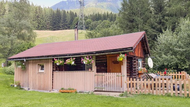 A wooden cabin with a red roof, surrounded by a wooden fence and lush greenery.