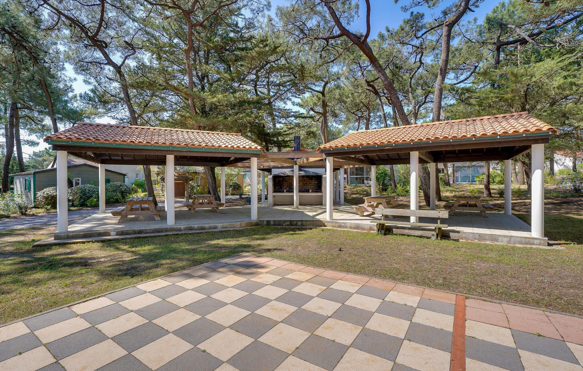 A gazebo with a tiled roof, surrounded by picnic tables and benches, set in a park with trees.