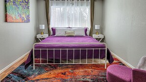 Un dormitorio con cama, ventilador de techo, ventana con cortinas, mesita de noche, lámpara, alfombra colorida y un cuadro en la pared.