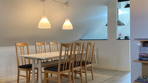 Ein Essbereich mit einem weißen Tisch, sechs Holzstühlen, zwei Pendelleuchten und einem Bücherregal mit Büchern.