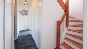 Un couloir avec un escalier en bois sur la droite, une porte au fond et un luminaire fixé au mur au-dessus.