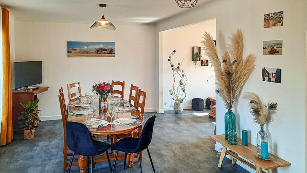 Un coin repas avec une table ronde dressée pour six personnes, entourée de chaises en bois. Un téléviseur est posé sur un meuble à gauche, et un vase rempli de fleurs se trouve sur la table.