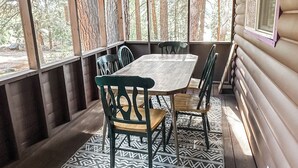 A covered outdoor seating area with a wooden table and chairs, a patterned rug, and a view of the surrounding forest.