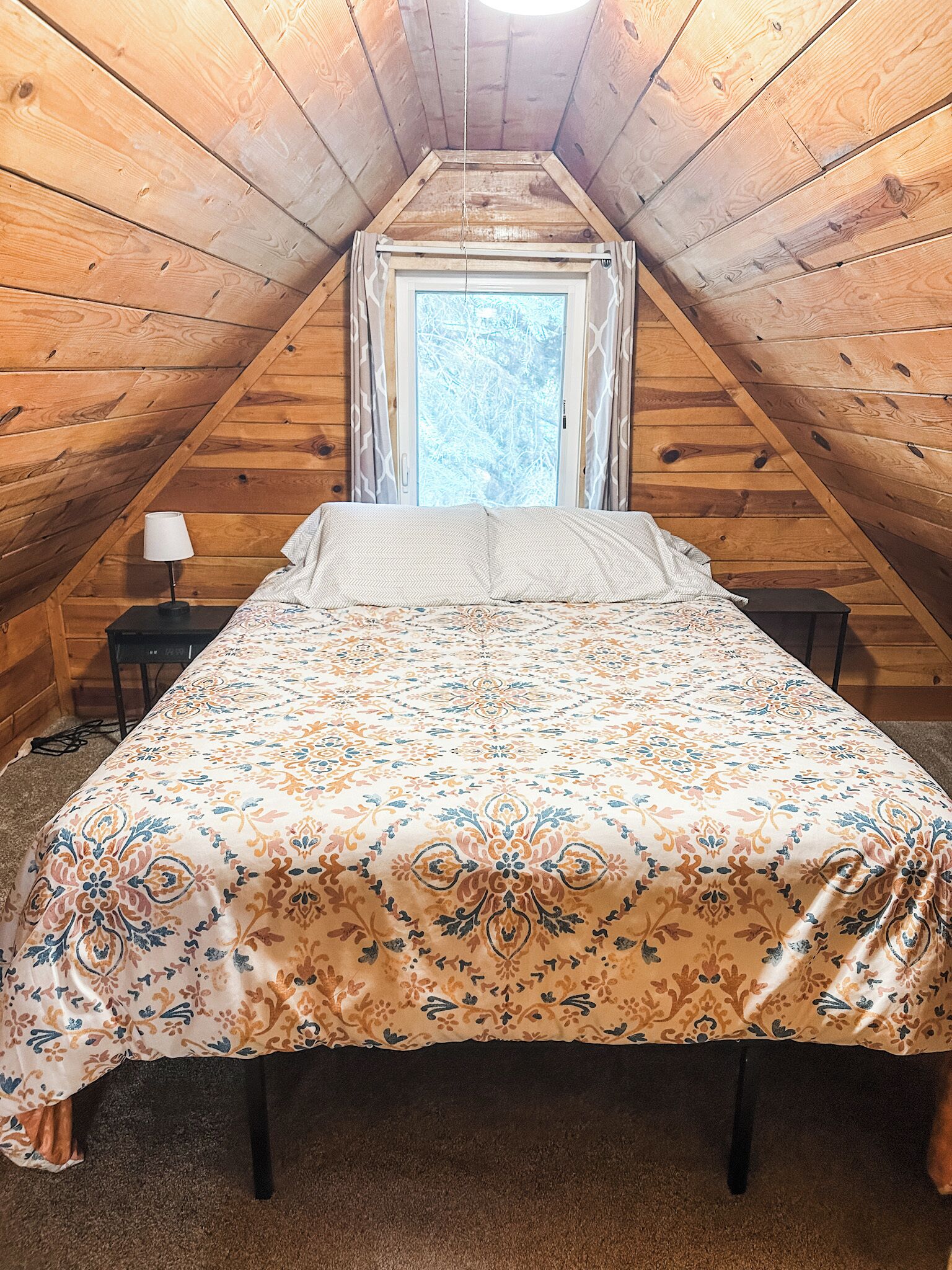 A bedroom with a sloped ceiling, a bed with a patterned bedspread, a nightstand with a lamp, and a window with curtains.