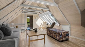 A spacious attic room with a sloped ceiling, wooden beams, a large window, a sofa, a table, and a colorful quilted ottoman. - 3 bedroom cozy home in Højslev (Højslev)