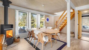 A modern living room with a fireplace, large windows, a dining area with wicker chairs, and a wooden staircase. - 3 bedroom cozy home in Højslev (Højslev)