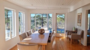 Un vaste espace de salle à manger avec une table et des chaises en bois, de grandes fenêtres offrant une vue sur les arbres et un lac, ainsi qu’un coin salon confortable avec une petite table et des chaises.