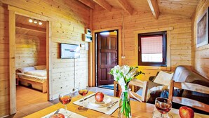 Interior de uma cabana de madeira com uma mesa de jantar posta para dois, uma cama, uma janela e uma porta.