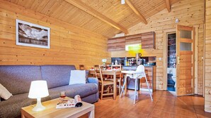 Interior acolhedor de uma cabana de madeira, com sala de estar que inclui um sofá, uma mesa de centro com abajur e uma mesa de jantar posta para dois. Uma cozinha com armários de madeira e uma área de refeições são visíveis ao fundo.
